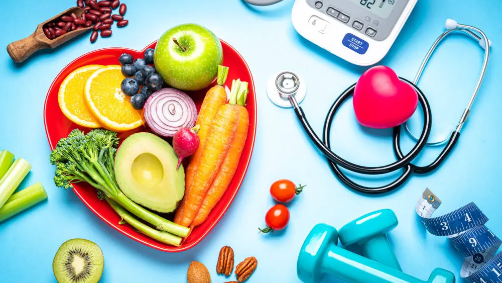 Healthy lifestyle concepts: red heart shape plate with fresh organic fruits and vegetables shot on blue background. A digital blood pressure monitor, doctor stethoscope, dumbbells and tape measure are beside the plate  This type of foods are rich in antioxidants and flavonoids that prevents heart diseases, lower cholesterol and help to keep a well balanced diet. High resolution 42Mp studio digital capture taken with SONY A7rII and Zeiss Batis 40mm F2.0 CF lens